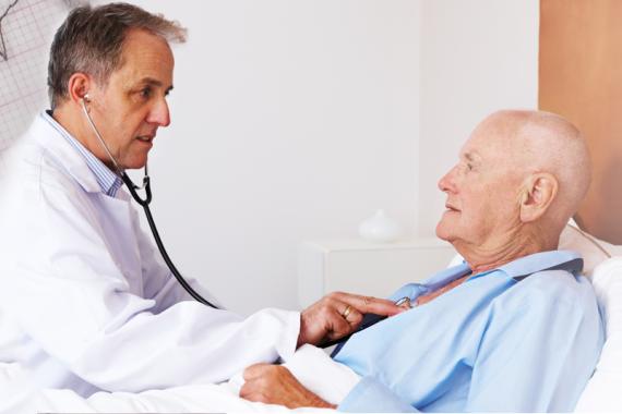 A doctor and a patient are in a hospital room. The patient is lying down and the doctor is holding a stethoscope on the patient’s chest